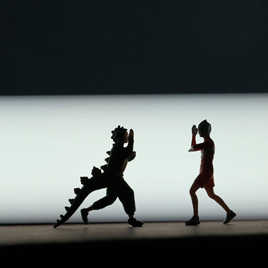 silhouettes of the green dinosaur-onesie monster and the gym-shorts hero facing off against a bright backlight, both tiny 1:64 scale figures