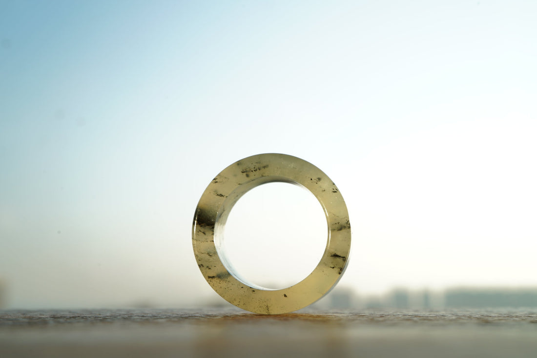 A translucent Xiuyu jade thumb ring standing on end on a ledge at dusk, backlit, with dark splashed-ink markings running through the stone.