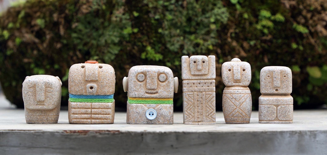 six small sandstone-coloured figures with simple carved faces standing in a row on a concrete ledge in front of moss