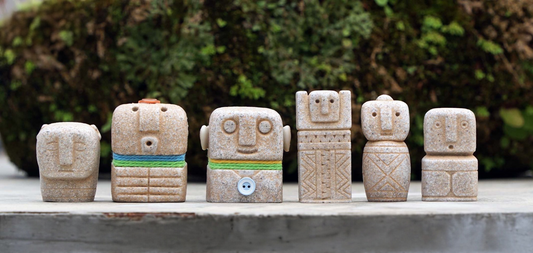 six small sandstone-coloured figures with simple carved faces standing in a row on a concrete ledge in front of moss