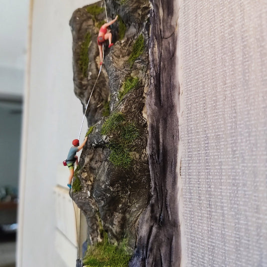 the pair of 1:64 rock climbers on a mossy miniature cliff, the woman in a pink top leading near the top, the man in an orange helmet below on the rope