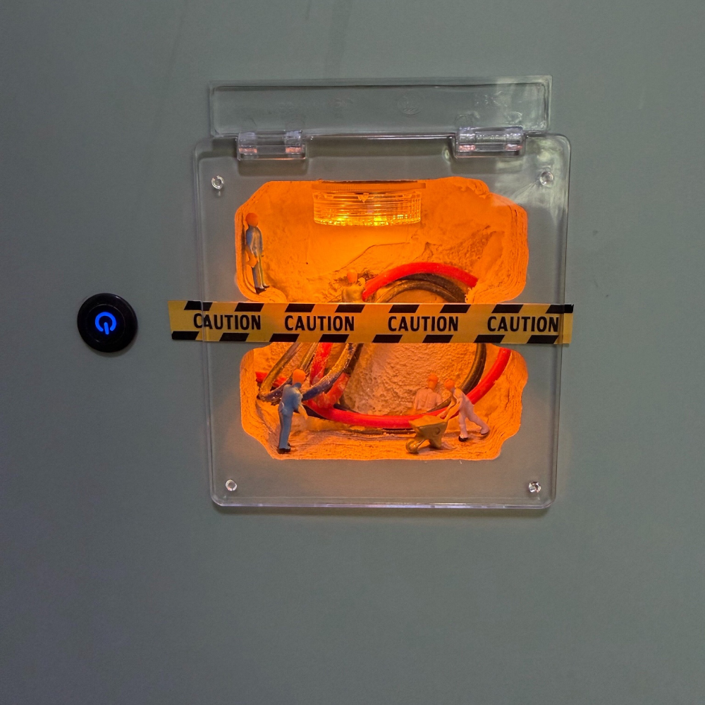the finished piece on a grey wall next to a light switch: a small glowing job site under a clear hinged cover