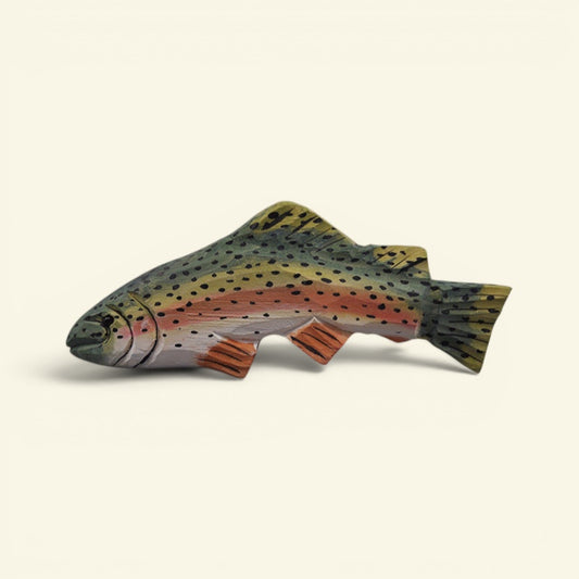 A handcrafted wooden fish carving shaped like a rainbow trout, featuring detailed natural wood grain, black spots, and orange-red accents, set against a solid off-white background. uggaz