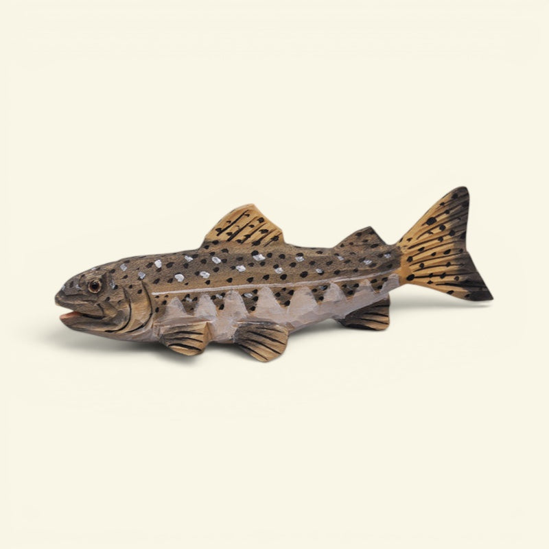 A carved wooden fish figurine, resembling a trout with brown and black spotted patterns and a warm tan finish, set against a solid light beige background. uggaz