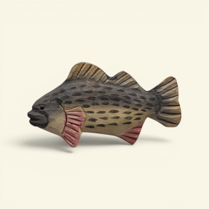 A hand-carved wooden fish figurine, likely representing a trout or salmon, featuring natural wood grain, dark spots, and pinkish-red gills and fins, set against a plain cream background. uggaz
