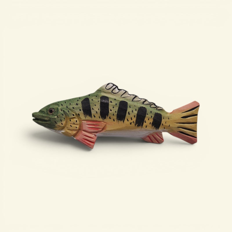 A hand-painted wooden fish figurine resembling a perch, featuring green and yellow-brown scales, black vertical stripes, and pinkish-red fins, set against a plain cream background. uggaz