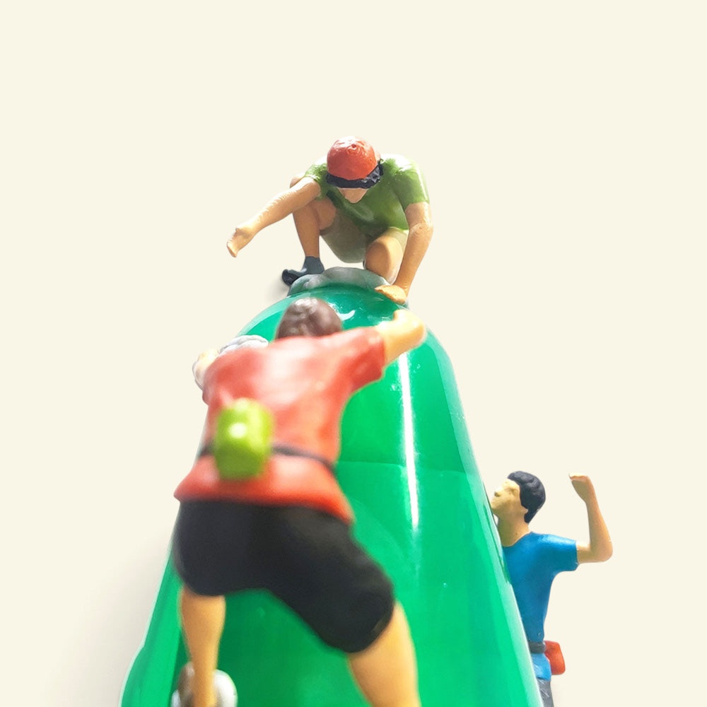 close-up from behind a 1:64 climber in a red shirt and black shorts with a green chalk bag, the green-shirt climber crouched on top, the blue climber on the right