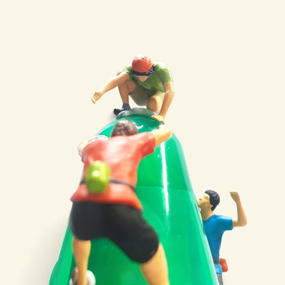 close-up from behind a 1:64 climber in a red shirt and black shorts with a green chalk bag, the green-shirt climber crouched on top, the blue climber on the right