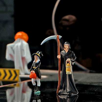 the 1:64 reaper with his scythe and lantern next to the little witch with her pumpkin bucket, the pumpkin-head ghost blurred behind them on a dark miniature street