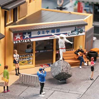 wide shot of a 1:64 street diorama: the happy astronaut on a rock in front of a little shop, tiny tourists with cameras, a guitarist on a bench