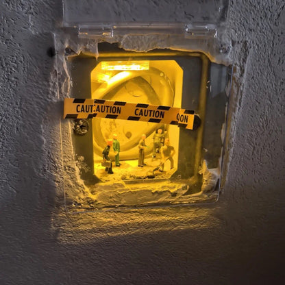 clear cover on a textured wall, a lit switch box behind it with caution tape and a few 1:87 workers