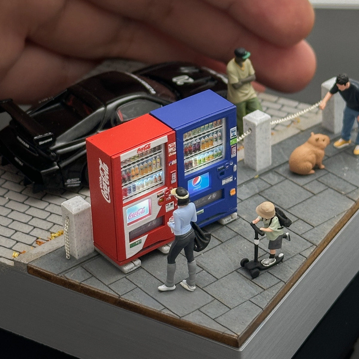 red and blue 1:64 vending machines on a miniature street corner with a real hand above for size; the figures, capybara, car and street are display pieces, not included