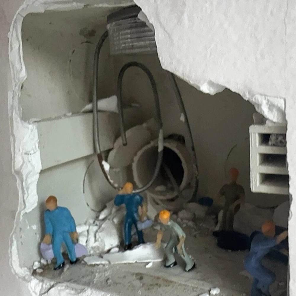 unlit wall hole with broken plaster, 1:87 workers in blue and grey clearing rubble around a pipe