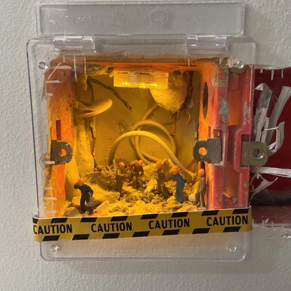 old switch box under a clear hinged cover, tiny workers digging in rubble beneath a warm led light, caution tape along the bottom