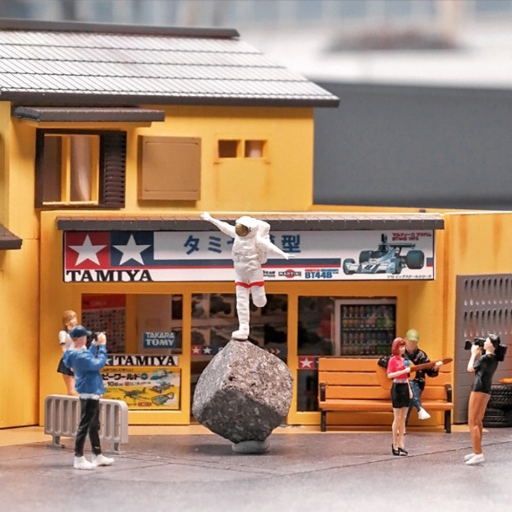 the happy astronaut doing his airplane pose on a boulder outside a 1:64 model shop diorama while tiny people take his picture