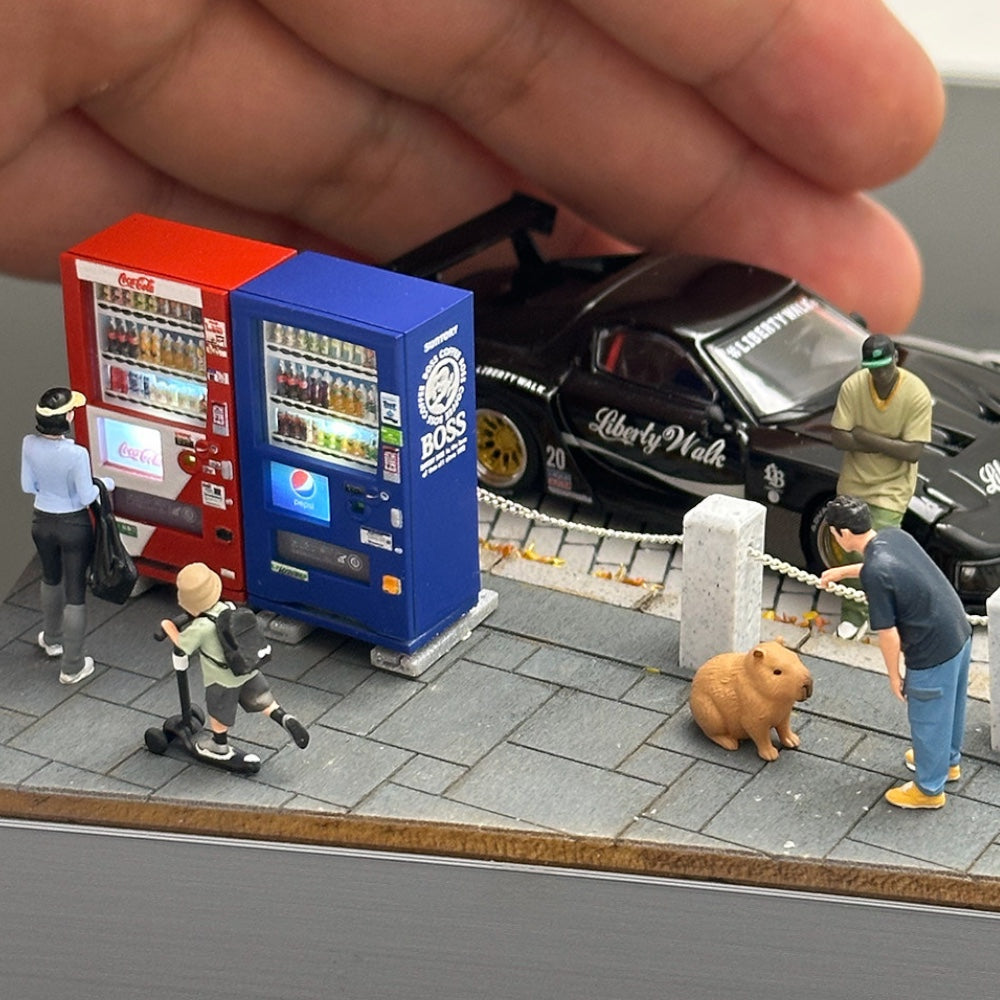 blue 1:64 vending machine next to the red one on a miniature cobbled street, a black diecast car parked behind; car, figures and street not included