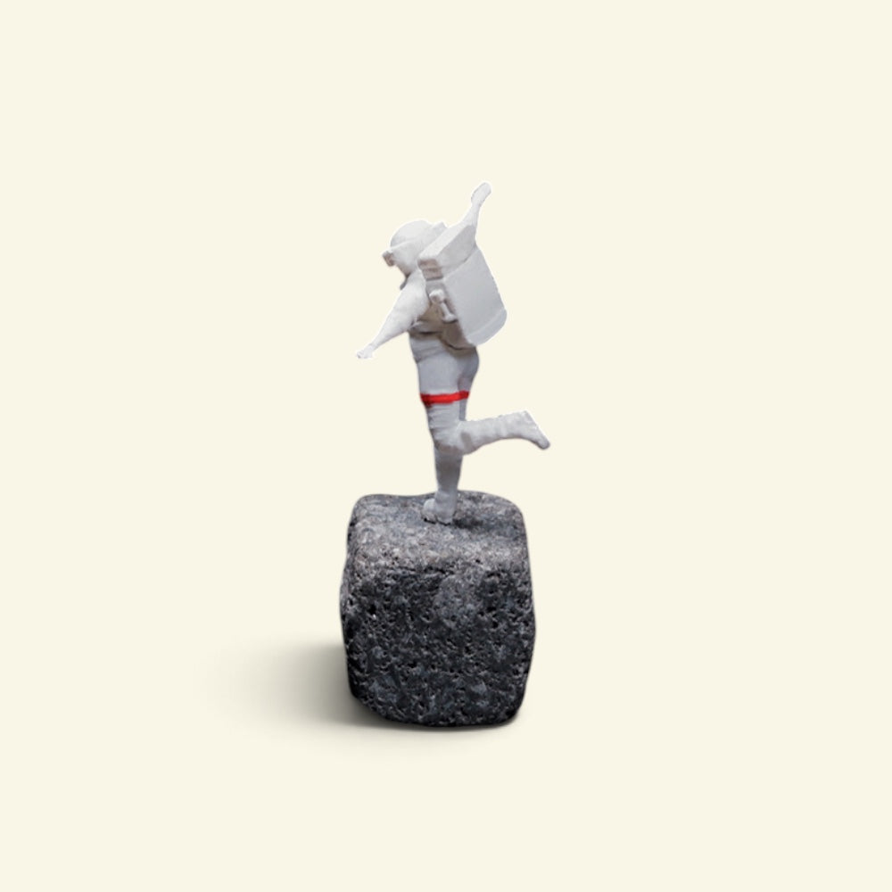 back view of the happy astronaut figurine on a rock, showing his life-support backpack and the red band around one leg