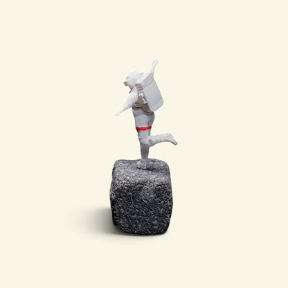 back view of the happy astronaut figurine on a rock, showing his life-support backpack and the red band around one leg