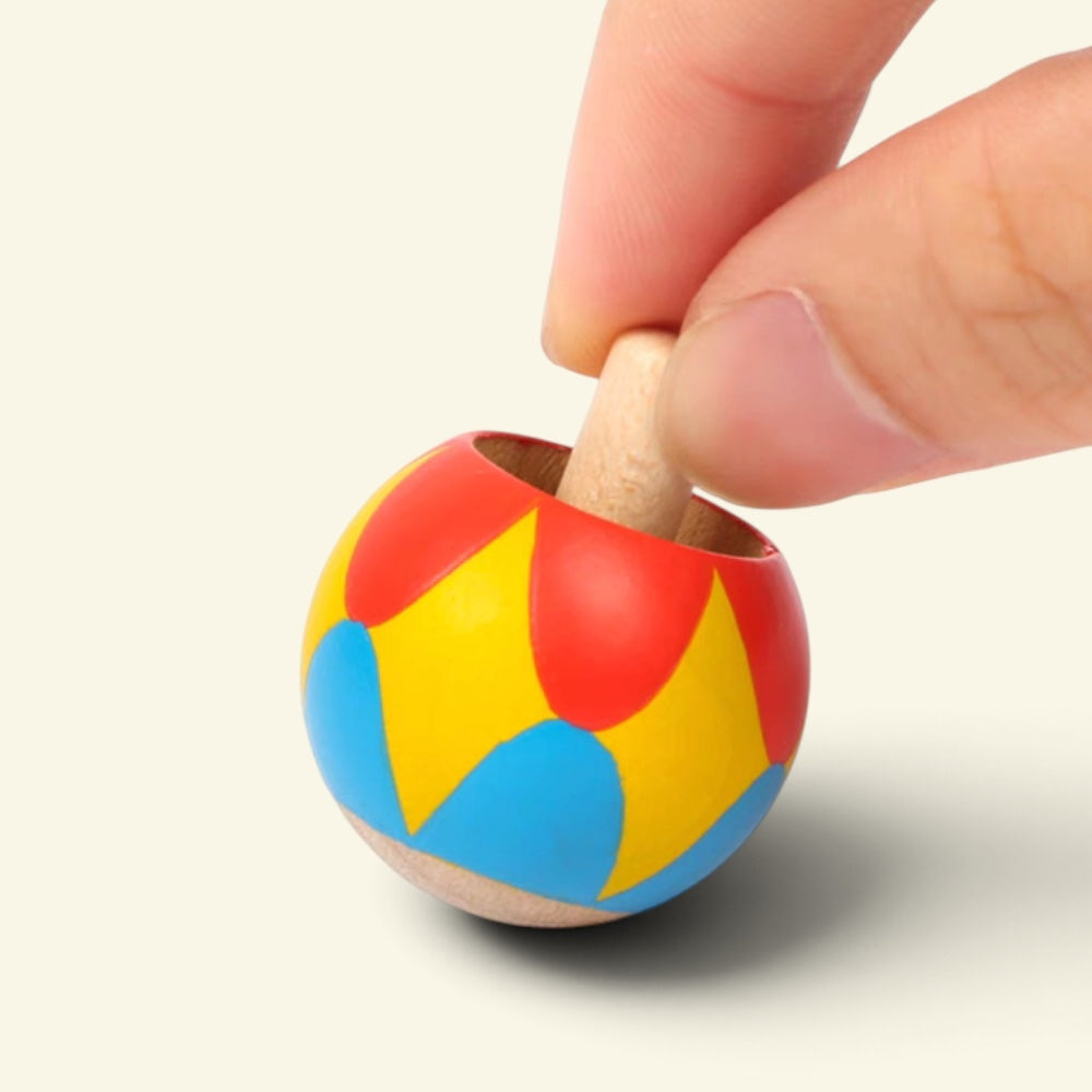 fingers holding the harlequin wooden tippe top by its stem, ready to twist and spin it, red, yellow and blue petals painted on the ball