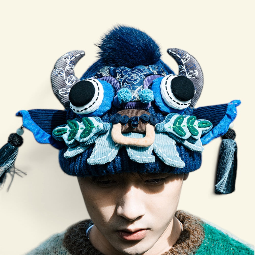 the ox head beanie in blue, close-up on a plain background — silver embroidered horns, black bead eyes, teal pompoms, and a wooden toggle nose, uggaz
