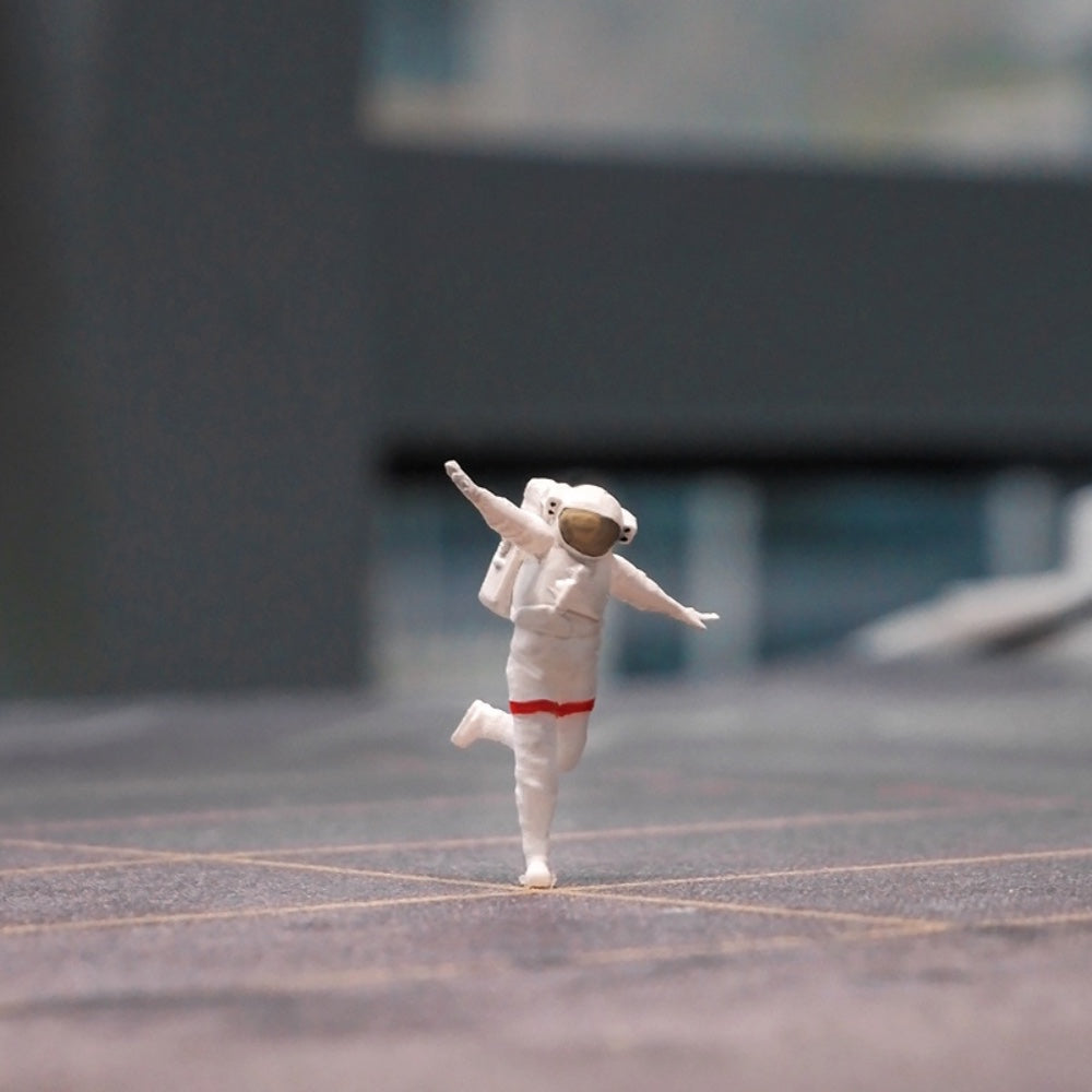 the happy astronaut 1:64 figurine dancing on one foot across a grey miniature pavement, city blurred behind him
