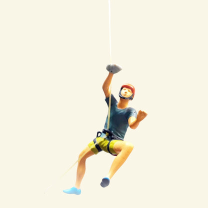 the orange helmet climber: a 1:64 hand-painted man in a dark grey t-shirt, yellow shorts and a harness, hanging on a thread and reaching up (the grey hold is a photo prop)