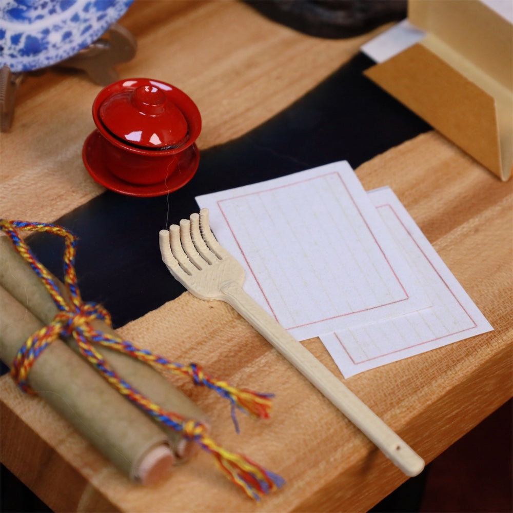 the tiny back scratcher on a miniature wooden desk beside tiny scrolls, a red lidded teacup and sheets of ruled paper, uggaz