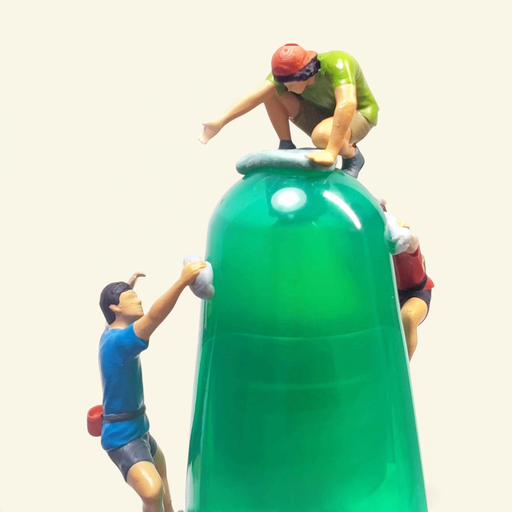 three tiny 1:64 scale hand-painted rock climbers on a green prop wall: one in a green shirt crouched on top, one in blue reaching for a hold, one in red on the side