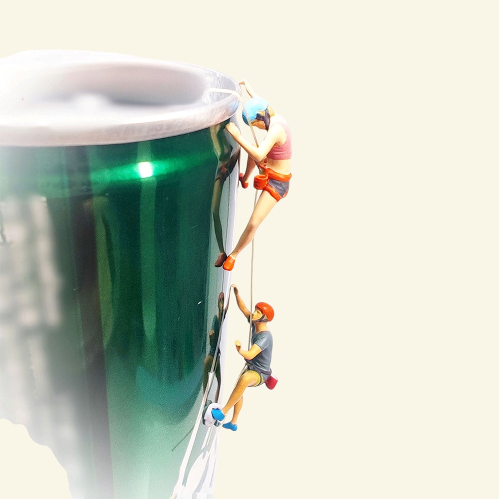 two tiny 1:64 scale hand-painted rock climbers roped up the side of a green drink can used as a prop wall: a woman in a blue helmet above, a man in an orange helmet below