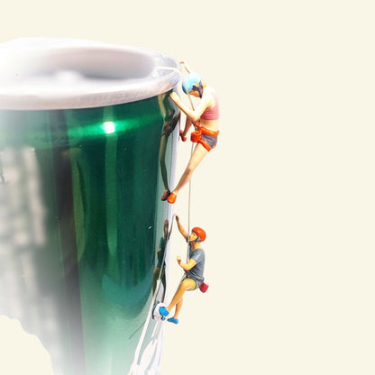 two tiny 1:64 scale hand-painted rock climbers roped up the side of a green drink can used as a prop wall: a woman in a blue helmet above, a man in an orange helmet below