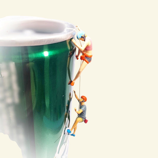 two tiny 1:64 scale hand-painted rock climbers roped up the side of a green drink can used as a prop wall: a woman in a blue helmet above, a man in an orange helmet below