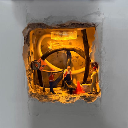 rough hole in a white wall lit from inside, tiny workers with a wheelbarrow, traffic cone and road sign among the cables