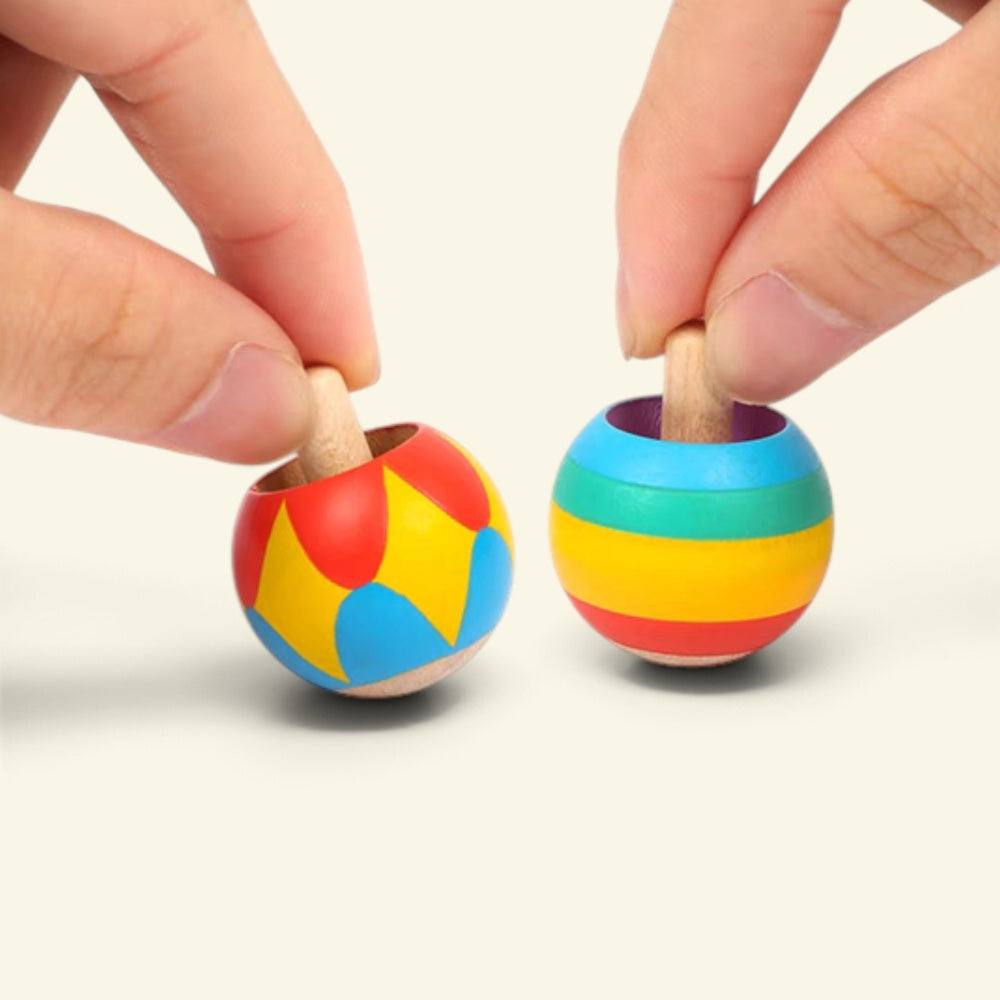 two hands each pinching a tippe top by its stem, harlequin on the left and rainbow on the right, about to spin them on a cream table