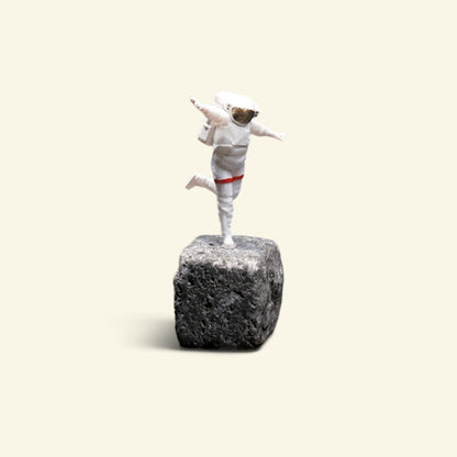 the happy astronaut, a 1:64 hand-painted resin figurine, balancing on one foot on a small grey rock with his arms out like wings — white spacesuit, gold visor