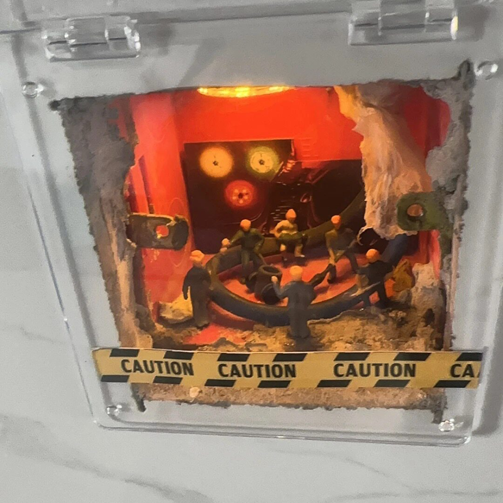 red-painted wall hole turned into a tiny control room, 1:87 workers gathered around a pipe behind a clear cover and caution tape