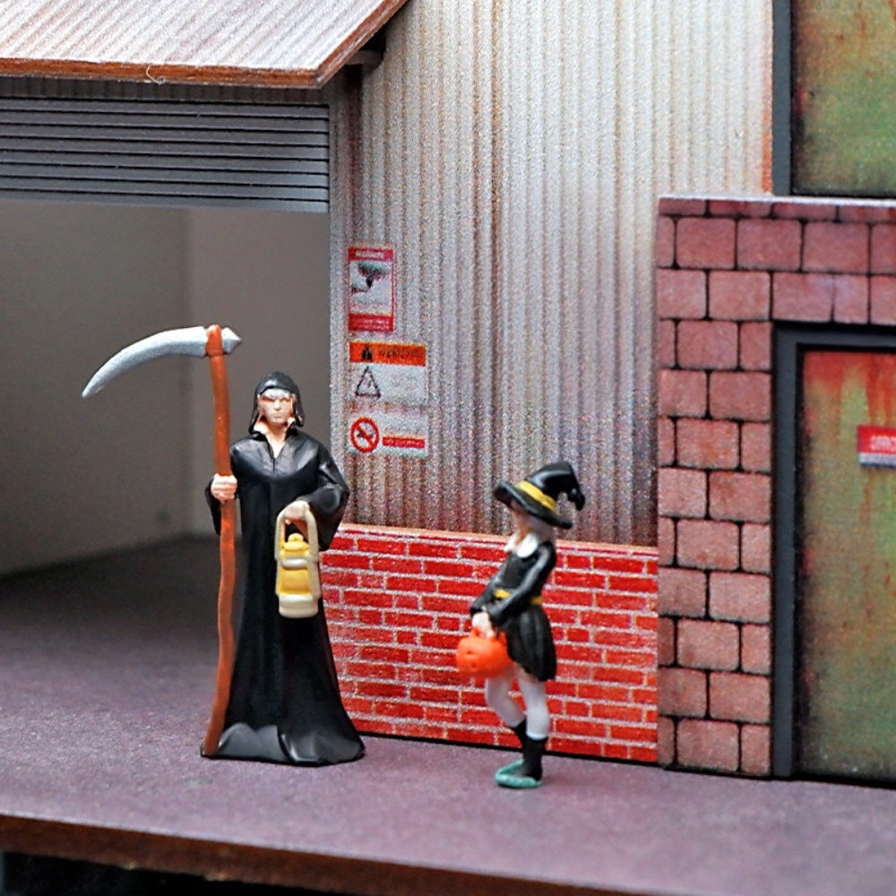 the 1:64 reaper and the little witch outside a miniature warehouse with a brick wall and a green door