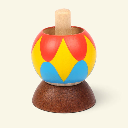 wooden tippe top painted with red, yellow and blue petal shapes, natural wood stem pointing up, sitting on a dark wood half-dome stand on a cream background