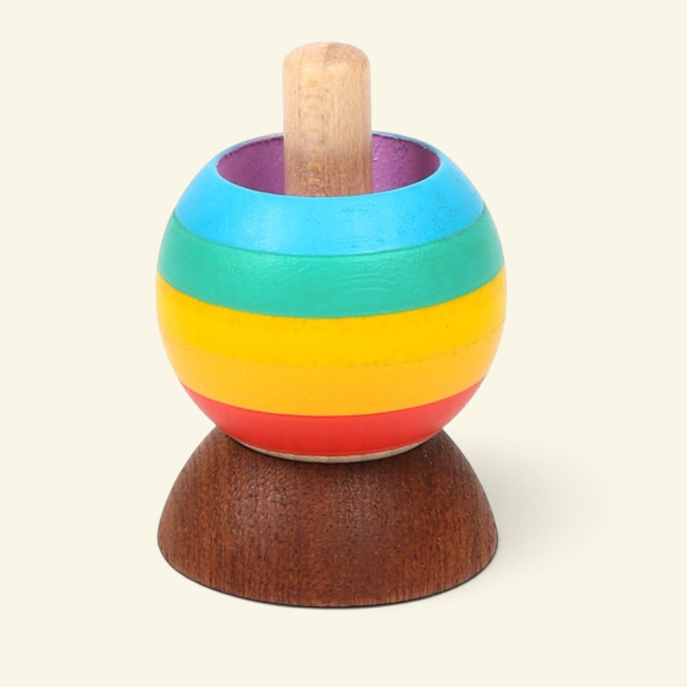 wooden tippe top painted in rainbow stripes — red, yellow, green and blue — purple inside the rim, natural wood stem, sitting on a dark wood stand